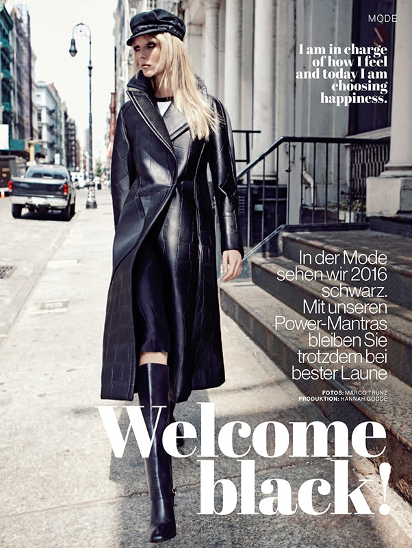 Fashion editorial on city street: Woman in black leather trench coat, knee-high boots, and cap. Buildings and vehicles line the background.