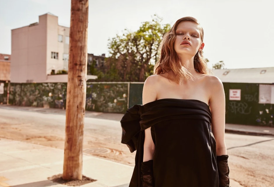 Fashionable adult female model wearing black dress and gloves, stands in urban street setting with buildings and wooden pole in the scene.