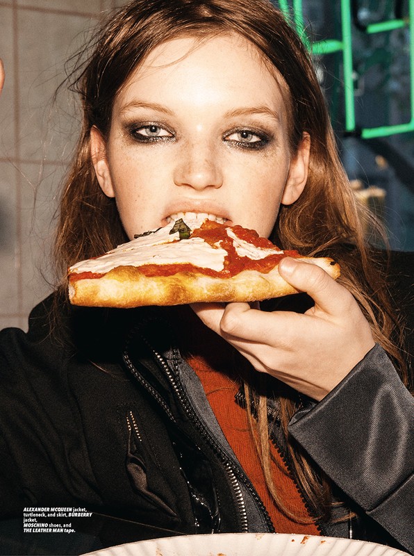 Young woman in black jacket eating a slice of pizza with tomato sauce and herbs, lit by a neon sign in the background.