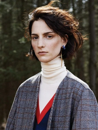 Portrait of a woman with short dark hair wearing a plaid coat, white turtleneck, and gold hoop earrings outdoors.