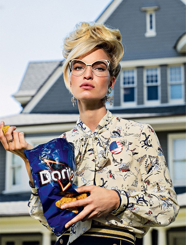 Woman in patterned blouse and glasses holds a bag of Doritos against an exterior house background in a fashion style portrait.