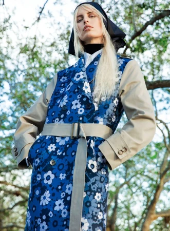 Model with blonde hair in a patterned blue floral trench coat with tan sleeves and a dark headscarf outdoors against tree branches.