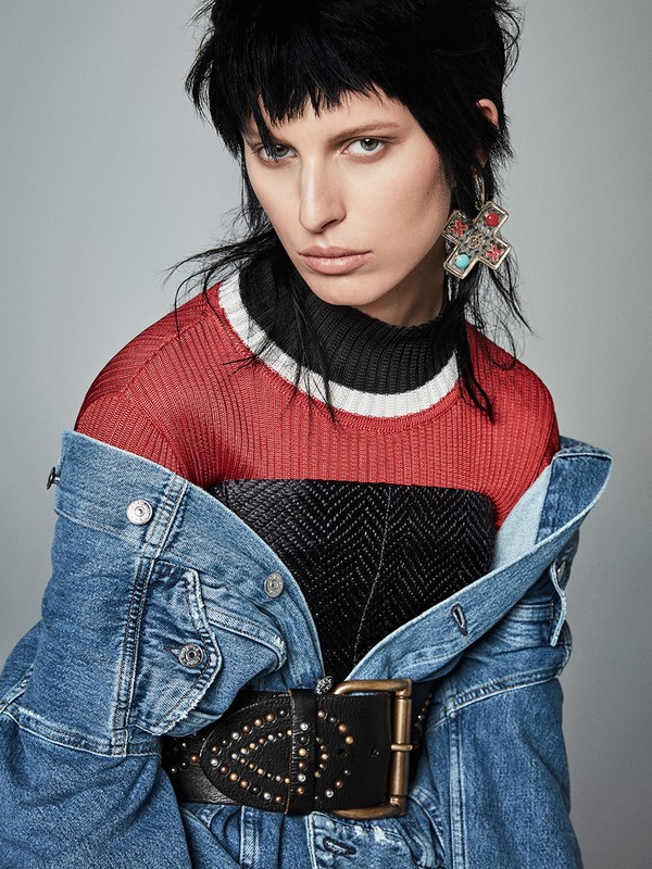 Close-up portrait of a woman with dark hair wearing a denim jacket over a red sweater and black top, accessorized with statement earrings.