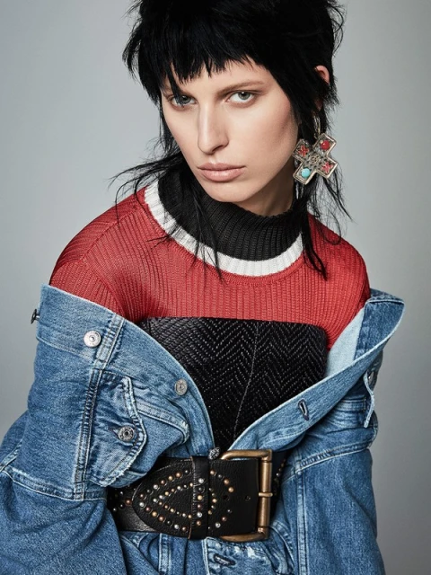 Close-up portrait of a woman with dark hair wearing a denim jacket over a red sweater and black top, accessorized with statement earrings.