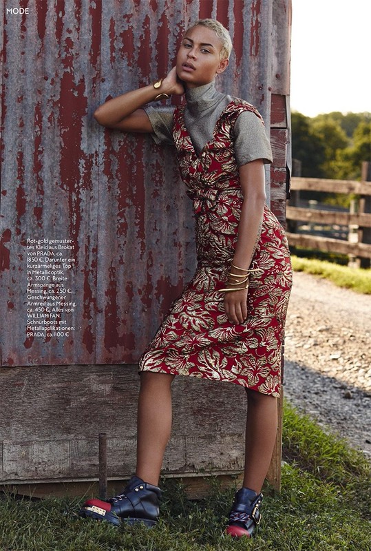 Adult female poses next to weathered metal wall outdoors, wearing patterned dress, metallic top, and platform shoes in a fashion editorial shot.