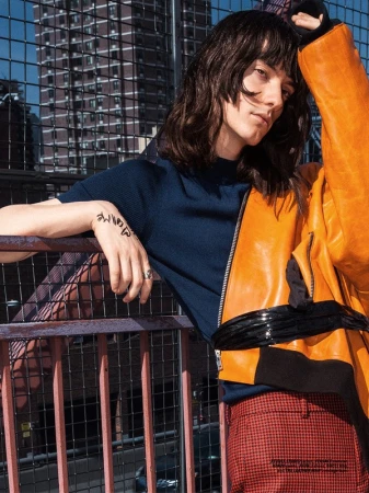 Model with dark shaggy hair wearing a navy top and orange jacket leaning against a pink railing outdoors.