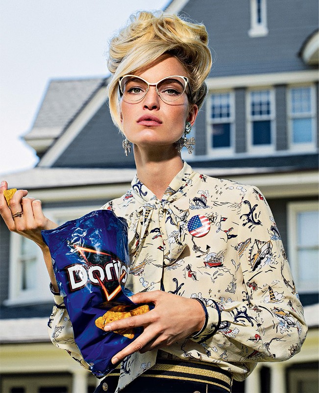 Adult woman wearing glasses and patterned blouse holds open a bag of Doritos in front of a residential house with blue siding.
