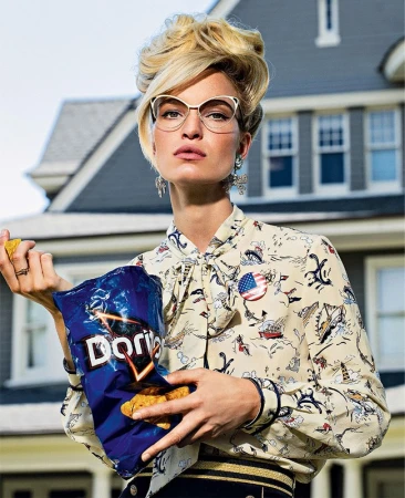Adult woman wearing glasses and patterned blouse holds open a bag of Doritos in front of a residential house with blue siding.