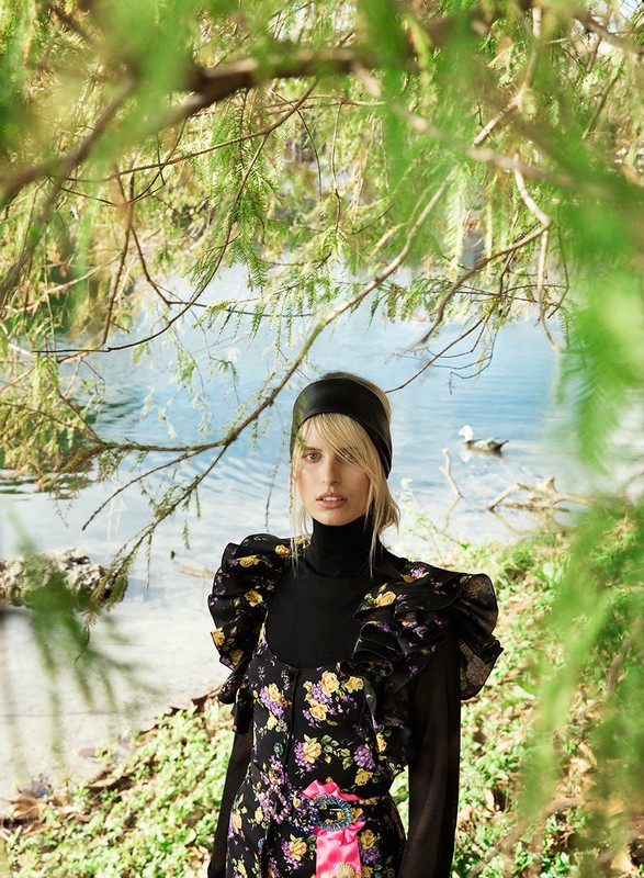 Woman poses in floral dress by a lake framed by foliage. Portrait of female model outdoors with water and greenery in the background.