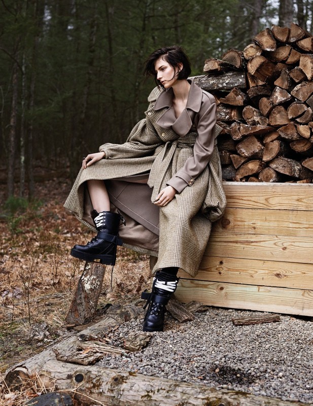 An adult woman sits on a wood box in a forest setting, wearing a long trench coat and black boots, with a woodpile behind her.