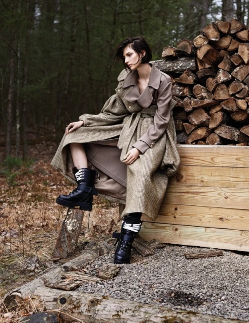An adult woman sits on a wood box in a forest setting, wearing a long trench coat and black boots, with a woodpile behind her.