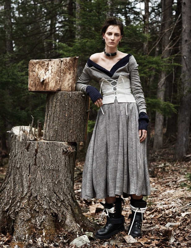 Female model in a wool dress, corset and boots poses next to a tree stump in a forest environment, fashion editorial photography.