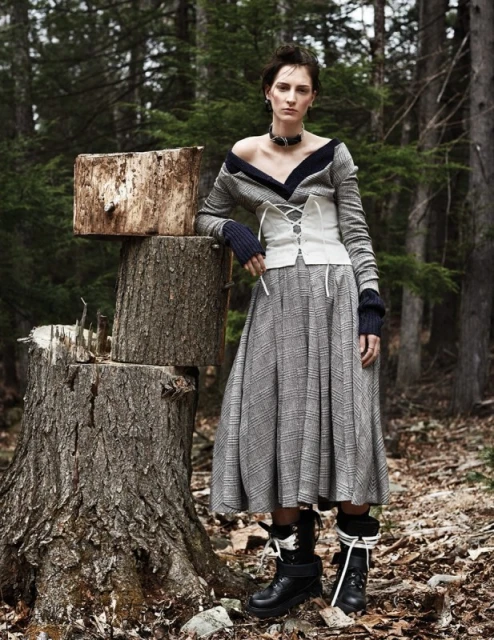 Female model in a wool dress, corset and boots poses next to a tree stump in a forest environment, fashion editorial photography.