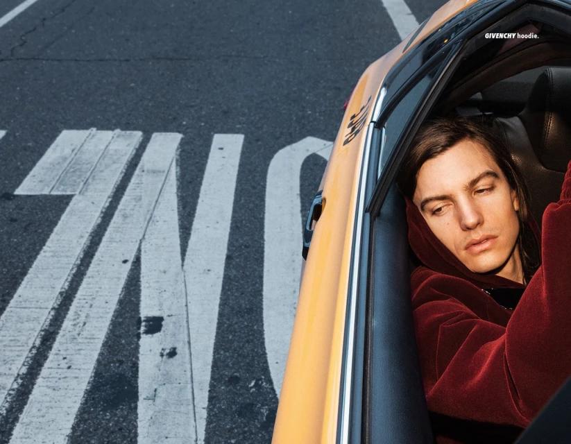 Adult male in a taxi with a burgundy hoodie, looking to the side with an urban street view through the front window. 