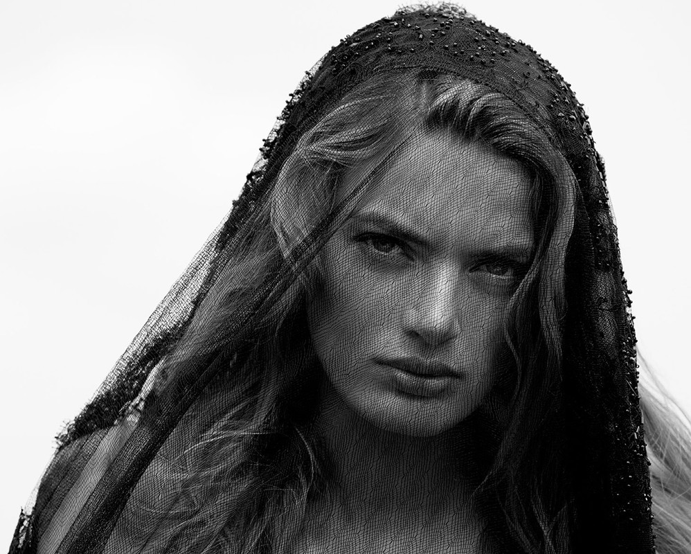 Black and white portrait of a blonde woman wearing a beaded black veil against a plain white background in a studio setting.