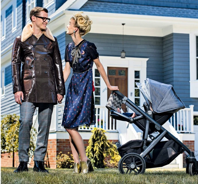 Retro-styled couple stands on a lawn with a stroller in front of a light-blue house. The woman wears a dress, and the man wears a leather coat.