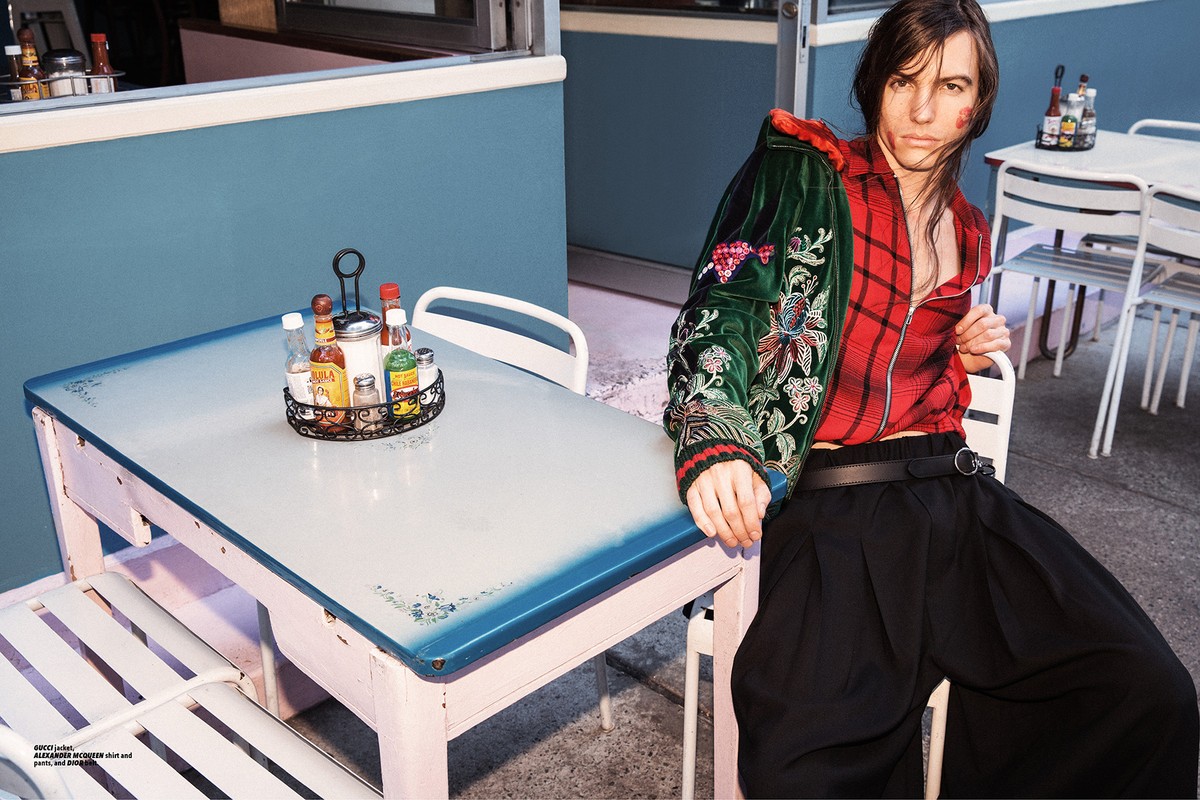 A model poses outdoors wearing a green embroidered jacket and red plaid jacket, paired with black pants, sitting at a diner table.