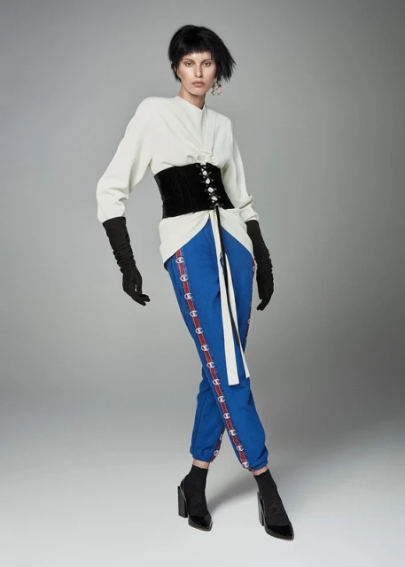 A full-length portrait of a young woman in a modern outfit, featuring a corset, sweater, and blue track pants, set against a grey backdrop.