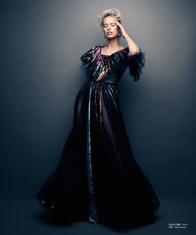 Fashion editorial shot of a blonde female model in a black sequined gown with colorful accents, posing in a studio setting against a dark gray backdrop.