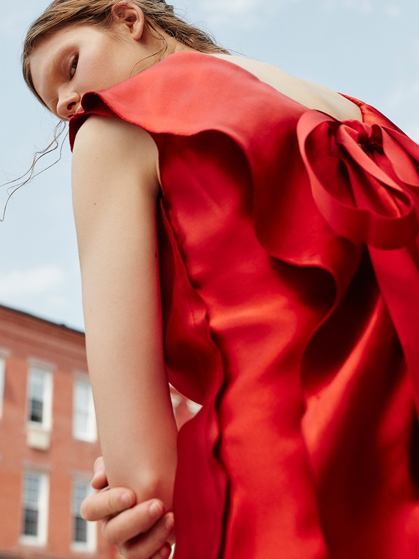 Woman in a red satin dress poses in an urban setting with a brick building, photographed from a low angle against a clear sky backdrop.