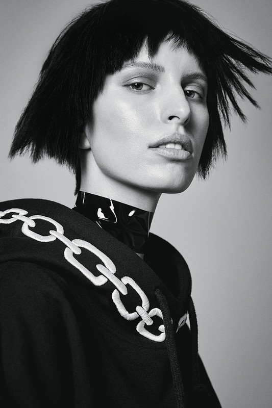 Portrait of a woman with short black hair wearing a hoodie with a chain design and a glossy black collar against a grey background.