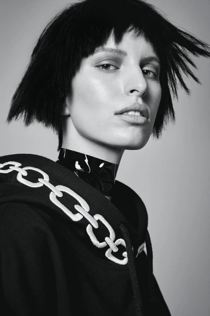 Portrait of a woman with short black hair wearing a hoodie with a chain design and a glossy black collar against a grey background.