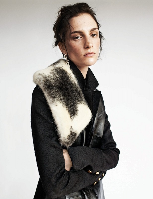 Woman in a dark coat and fur stole poses in a studio setting. The portrait captures a minimalist fashion aesthetic with neutral backdrop.