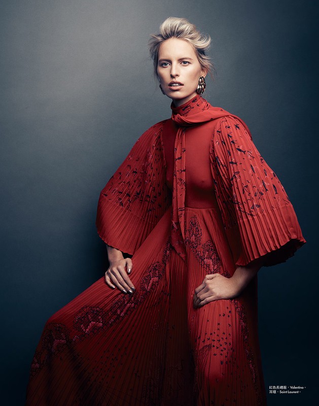 A blond woman wearing a pleated red dress with bishop sleeves posing against a dark gradient backdrop.