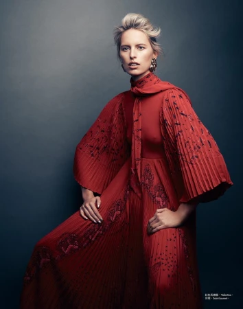A blond woman wearing a pleated red dress with bishop sleeves posing against a dark gradient backdrop.