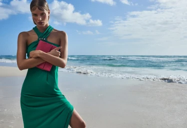 Model in green halterneck dress holding a pink clutch on a sandy beach