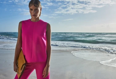 Model in vibrant pink sleeveless top holding a mustard yellow clutch on a sandy beach