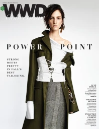 Model wearing an olive green coat, white laced corset, and grey skirt against a white studio background with magazine cover text.