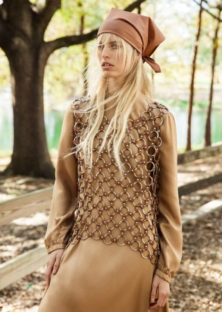 Model with long blonde hair wearing a brown headscarf and ring-mail overlay dress outdoors near trees