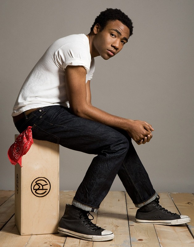 Portrait of an adult male seated on a wooden box, wearing a white t-shirt, dark jeans, and black high-top sneakers against a grey background.