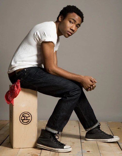 Portrait of an adult male seated on a wooden box, wearing a white t-shirt, dark jeans, and black high-top sneakers against a grey background.