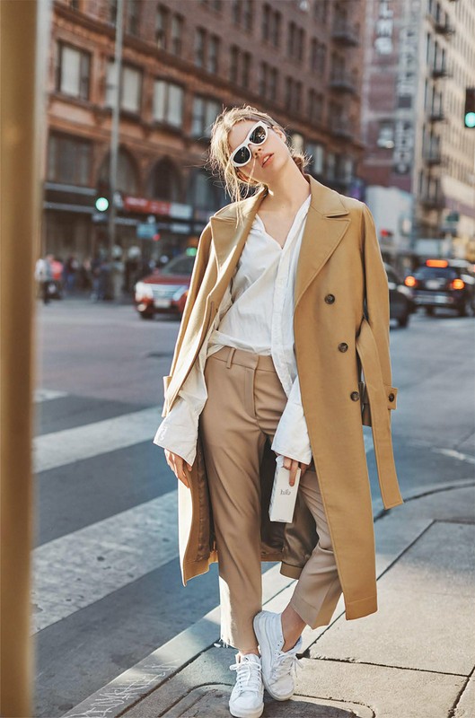 A stylish woman in a trench coat and sunglasses poses on a city street, blending fashion with an urban environment and pedestrian crossing.