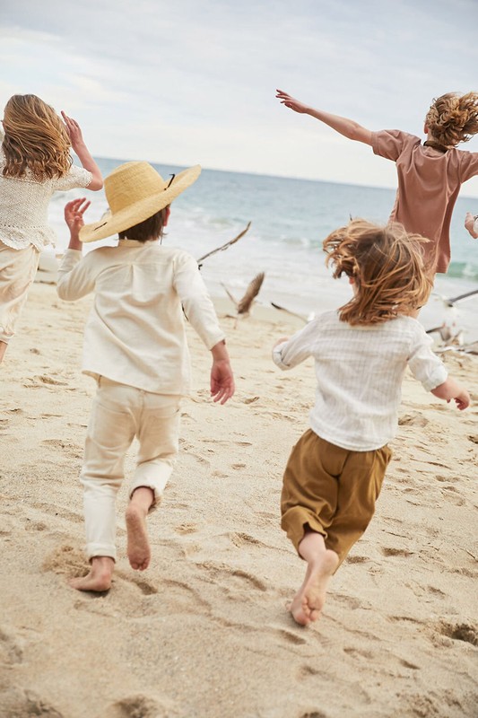 Four children run barefoot on a sandy beach toward the ocean under an overcast sky, wearing light-colored linen clothing and a straw hat.