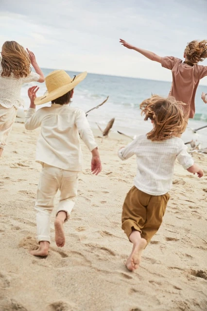 Four children run barefoot on a sandy beach toward the ocean under an overcast sky, wearing light-colored linen clothing and a straw hat.