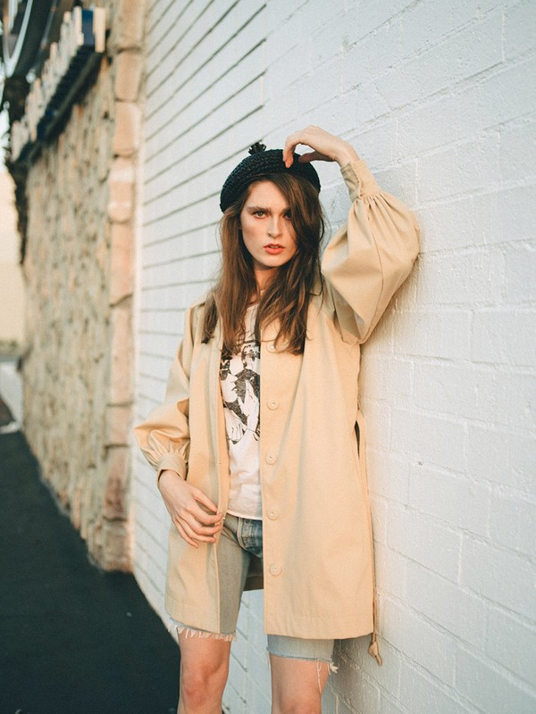 A young adult woman with long brown hair and a black beret poses against a white brick wall in a beige coat, with a concrete wall in the background.