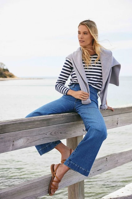 Blonde woman in jeans and striped top sits on a wooden railing with water and land visible in the background, styled casually.