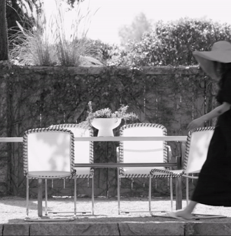 A woman with a wide-brimmed hat walks past a garden table and chairs on a stone patio, with greenery in the background in black and white.