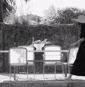 A woman with a wide-brimmed hat walks past a garden table and chairs on a stone patio, with greenery in the background in black and white.