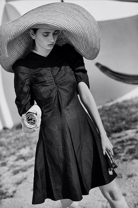 A woman in a black dress and wide-brimmed hat poses on a grassy lawn with a white umbrella, holding a rolled newspaper.