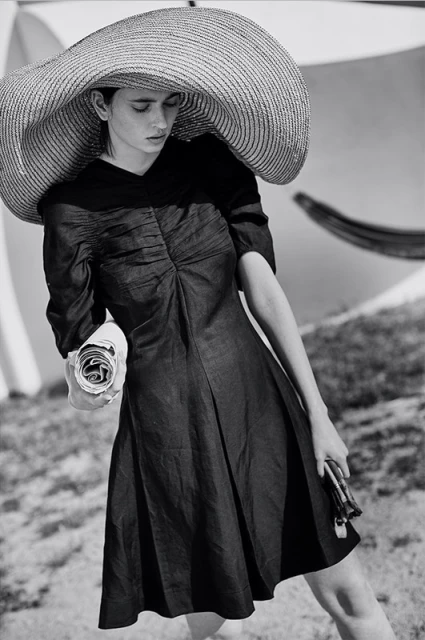 A woman in a black dress and wide-brimmed hat poses on a grassy lawn with a white umbrella, holding a rolled newspaper.