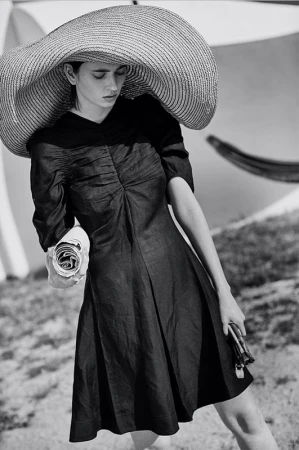 A woman in a black dress and wide-brimmed hat poses on a grassy lawn with a white umbrella, holding a rolled newspaper.