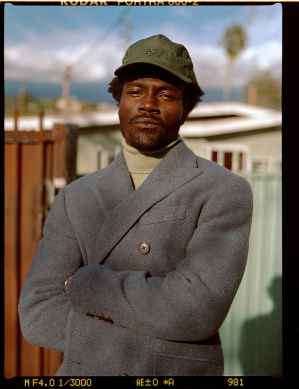 Black man wearing a grey double-breasted blazer, green cap, and turtleneck outdoors in Los Angeles with arms crossed.