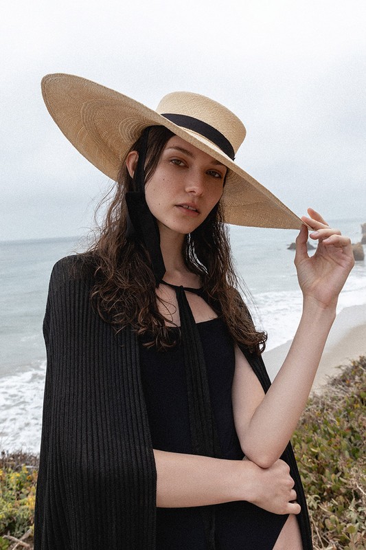 Woman in black swimwear and cape posing on beach with ocean background, wearing wide-brimmed straw hat in an eye-level shot.