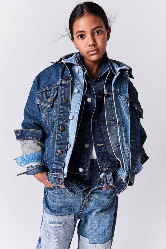 Child model poses in layered denim jackets and patchwork jeans against a white backdrop in a fashion forward editorial studio shot.