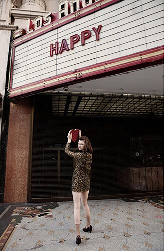Young woman holding a heart-shaped box standing in front of the Los Angeles Theatre with the word 'Happy' on the marquee.