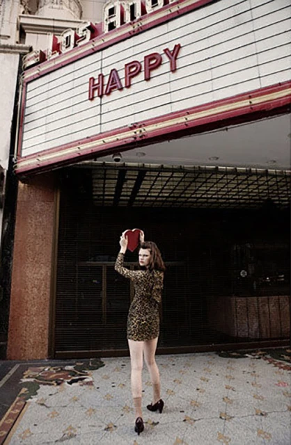 Young woman holding a heart-shaped box standing in front of the Los Angeles Theatre with the word 'Happy' on the marquee.